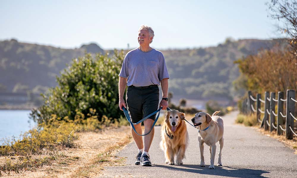 John walking two dogs on a walking trail by a lake and mountains